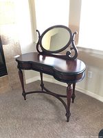 Full frontal shot of wooden vanity table with kidney-shaped top, three drawers, oval swivel mirror, and spindle legs.
