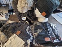 Four black winter toques including two fleece-lined floppy style and two fuzzy water-repellant with ear flaps and face masks, all Bluetooth enabled with built-in speakers. Items shown on mannequin head and packaged on a table.
