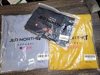 Three wrapped Jed North Apparel clothing items: blue ribbed dress, yellow ribbed dress, and black tank top in packaging.