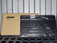 Arteck HB030B Universal Backlit Bluetooth Keyboard with protective plastic wrap and original packaging box displayed on a metal surface.