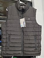 Front view of the black Venustas men's medium electrically heated padded vest on a hanger showing the front zipper, zippered pockets, and Venustas logo with power button.