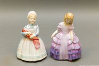 Front view of two Royal Doulton figurines showing 'Rose' with bonnet and apron holding red cloth, and 'The Rag Doll' in pink-purple layered dress.