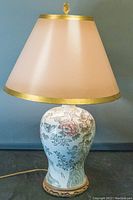 Front view of table lamp showing off-white tapered shade with gold trim and ceramic base with floral and bird designs.