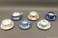 Six bone china teacup and saucer sets arranged in two rows, displaying varied floral patterns and colors including blue, pink, white, and green.