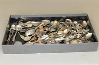 Overview of a metal tray filled with assorted collectible souvenir spoons of various designs and sizes.