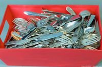Top view of orange bin filled with assorted flatware including forks, spoons and knives showing mixed metal finishes.
