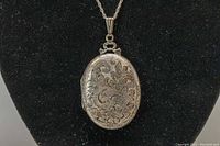 Front view of sterling silver oval locket pendant with detailed floral etching on black display stand.