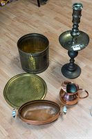 Brass pail, hammered brass tray, copper pitcher, tall brass candleholder, and oval copper pot with porcelain handles displayed on wood floor.