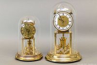 Two vintage brass mantle clocks under clear plastic domes; one larger 12 inch Kundo Ergo model, one smaller Kieninger & Obergfell Kundo clock. Both have brass bases and suspended rotating balls pendulums.