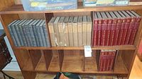 Books on wooden shelves showing three distinct sets: blue cloth 1922 Elbert Hubbard writings, beige cloth New Standard Encyclopedias with damage, and red 4th edition encyclopedia volumes.