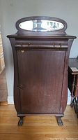 Front view of the antique wooden cabinet showing the upper drawer, single door, latch, and attached oval mirror.