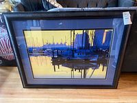 Framed art print showing a harbor with boats at sunset, reflections in water, dark blue matte, black frame.