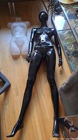 Black glossy full-body female mannequin lying on wooden floor next to white hanging torso form showing overall condition and scratched surface