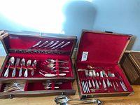 Both wooden boxes with silver plate flatware sets displayed open, showcasing contents of forks, knives, spoons, and serving utensils with red velvet lining and box exterior detail.