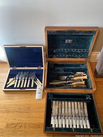 Photo showing three sets of silver plate flatware in three storage boxes of different styles and fabrics, viewed from above.