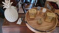 Two Equity brass clocks, small decorative brass pineapple, salt and pepper shakers, on a round wooden tray. Large white ceramic pineapple jar next to the tray. Pineapples show damage.