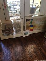 Full view of three framed artworks leaning against wall: large Bermuda farm house print, smaller colorful abstract piece, and tiny card-like art