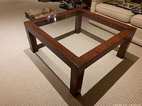 Square coffee table with wood frame and clear plexiglass top shown at an angle on a carpeted floor.