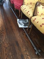 Full view of the curtain rod laid on the floor beside upholstered furniture, showing the length and overall condition of the rod with attached decorative finials.
