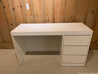 Front view of the white wood and laminate desk with three drawers on right side and open leg room left side, placed against wood-paneled wall and carpeted floor.