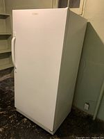 Frigidaire white vertical freezer showing front and right side in basement setting on worn floor.