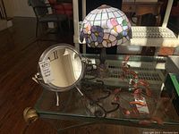 Photo showing Tiffany style lamp with stained glass-pattern plastic shade, round metal framed makeup mirror, metal wall coat rack, and copper leaf wall art on glass table.