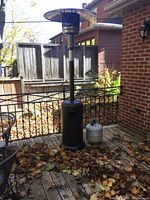 Full view of the outdoor patio heater standing on a wooden deck with a black metal fence background and autumn leaves around. The heater has the large reflector and propane tank attached, with a second propane tank placed nearby.