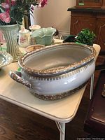 Photo showing a large oval ceramic planter with gold and brown ornate trim and handles on a white table.