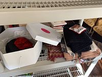 Overview of white box containing black hat and adjacent stack of scarves on shelf