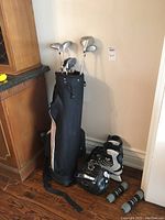 Wide view showing full golf bag with clubs standing upright beside the Bauer hockey helmet, Softtec skates, and Nike hand weights on wooden floor.