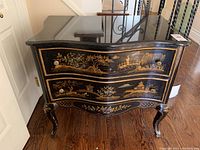 Front view of vintage wooden table with black finish, Asian-themed gold inlays on drawer fronts and floral motifs at base, marble top with curved edges.