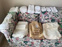 Full view of multiple folded white and beige tablecloths and cloth napkins displayed on floral couch.