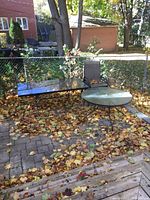 Photo showing one round outdoor patio table with glass top and one rectangular wrought iron patio table with glass top, four chairs partially visible, leaves on the ground.