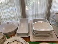 Set of white ceramic bakeware on table with curtain background. Large oval covered baker, several rectangular and square baking dishes stacked, large decorative serving tray underneath.