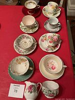 Full view of all 8 vintage cups and saucers arranged on red tablecloth, showing various floral designs and shapes
