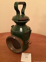 Front angle showing green metal body, carrying handle, and glass lens cover of the railway oil lamp