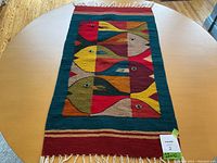 Overall view of the rectangular woven textile laid flat on a table, showing all eight stylized fish in bright colors and fringed ends.