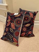 Two decorative pillows leaning against a wall on carpeted floor showing dark blue front with colorful floral and leaf embroidery.