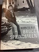 Front cover dust jacket showing young Bruce Springsteen leaning on a classic car on a suburban street with the title 'Born To Run Bruce Springsteen'.