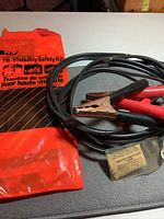 Image showing bright orange hi-visibility safety sleeve with black diagonal stripes and text, along with black and red booster cables with clamps coiled next to it. A small plastic sleeve attached to the cables is visible with instructions.