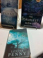 Three Louise Penny novels Still Life, Dead Cold, and The Cruelest Month shown with covers and standing display