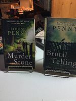 Two Louise Penny books shown upright on stands: 'The Murder Stone' and 'The Brutal Telling'