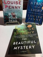 Photo showing three Louise Penny novels: Bury Your Dead, A Trick of the Light, The Beautiful Mystery, arranged standing and lying on a white surface.