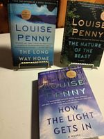 Three Louise Penny novels displayed upright and lying on a white surface showing titles and cover designs.