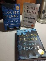 Three books by Louise Penny standing and lying on a table: 'A Great Reckoning' (standing left), 'Kingdom of the Blind' (standing right), and 'Glass Houses' (lying flat). Covers feature blue and white cover art with gold and white text.