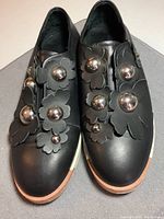 Pair of black leather shoes decorated with various sizes of silver spherical embellishments on top of leather flower appliques, showcasing front view and sole contrast.
