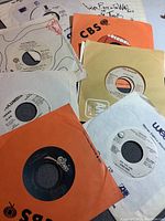 Photo showing a collection of vintage 45 RPM vinyl records in various paper sleeves including CBS, Columbia, Warner Bros., and Epic labels, arranged on a flat surface. Visible titles include Pink Floyd and John Lennon records.