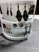 White earring rack holding six pairs of earrings: small sterling silver hoops, large black ornamented sterling silver dangles, small sterling silver curved dangles, blue long dangles, gold-toned Paua shell and mother-of-pearl studs, and gold-toned delicate dangles.