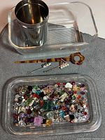 Top view of glass butter dish, metal tin holding rhinestones, and three hair pins including hair pick and two decorative pins, arranged on gray surface.