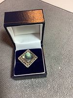 Small sterling silver square pin with paua shell inset in a blue velvet-lined jewelry box.